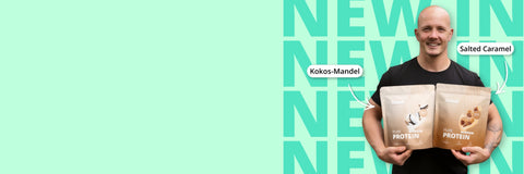 Der Mann hält zwei beige-farbene Proteinpulver-Beutel, die mit ’kokos-mandel’ und ’salted caramel’ beschriftet sind.