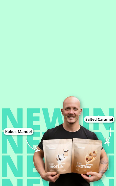 Zwei beige Beutel mit reinem Proteinpulver, einer beschriftet mit ’kokos-mandel’ und der andere mit ’salted caramel,’ gehalten von einem lächelnden Mann vor einem minzgrünen Hintergrund.