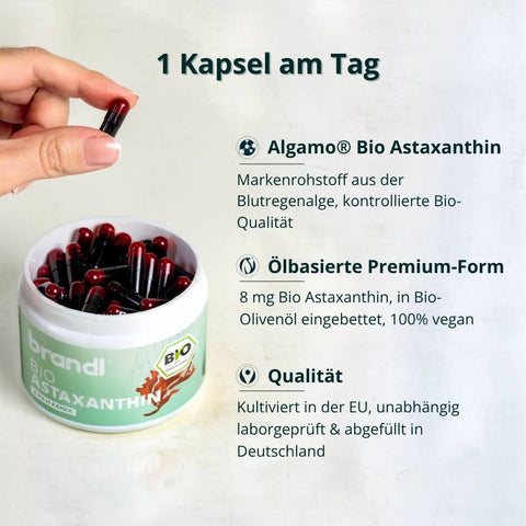 Eine hand hält eine schwarze und rote kapsel über einem weißen topf brandl bio astaxanthin mit vielen kapseln