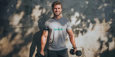 Ein Mann, der ein graues kurzärmeliges T-Shirt trägt, auf dem das Wort 'brandl' in Grün über der Brust gedruckt ist.