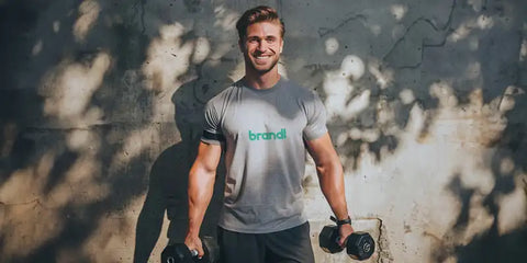 Mann steht mit brandl Shirt und Gewichten vor einer Wand
